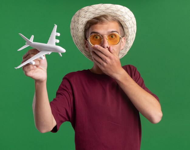 Scared young handsome guy wearing red shirt with glasses and hat holding toy airplane covered mouth with hand isolated on green wall