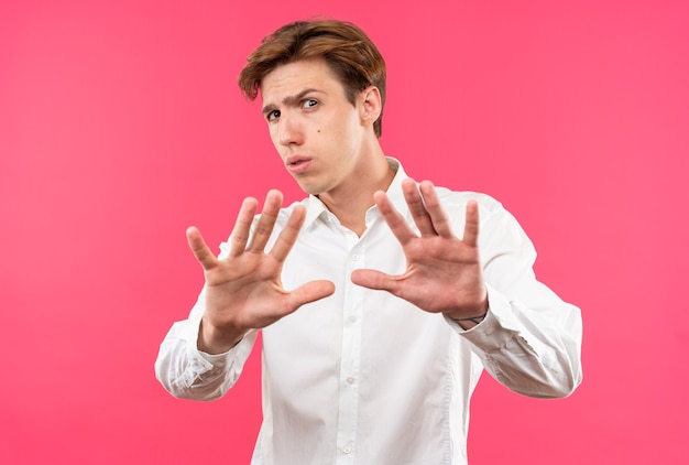 Scared young handsome guy wearing white shirt holding out hands at camera isolated on pink wall