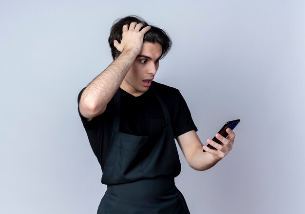 Scared young handsome male barber in uniform holding and looking at phone and grabbed head isolated on white