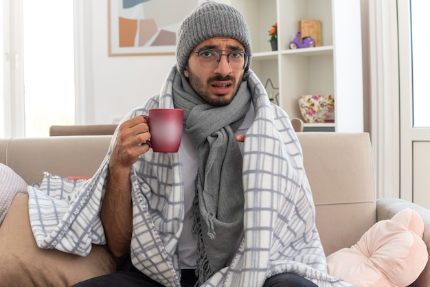 scared young ill caucasian man in optical glasses wrapped in plaid with scarf around his neck wearing winter hat holding and pointing at cup sitting on couch at living room