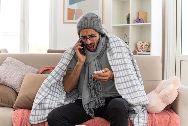 scared young ill caucasian man in optical glasses wrapped in plaid with scarf around his neck wearing winter hat talking on phone and looking at tissue sitting on couch at living room