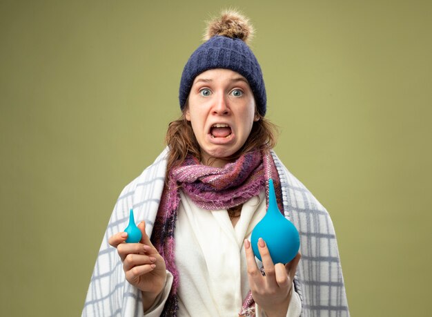 Scared young ill girl wearing white robe and winter hat with scarf wrapped in plaid holding enemas isolated on olive green