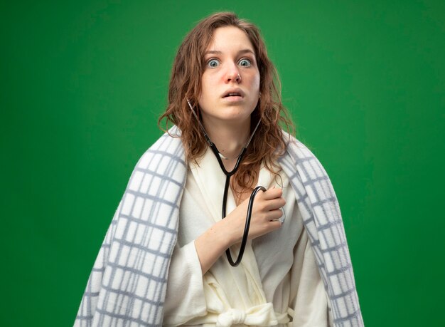 Scared young ill girl wearing white robe wrapped in plaid listening to her own heartbeat with stethoscope isolated on green