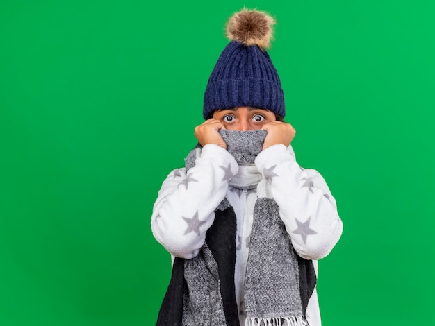 Scared young ill girl wearing winter hat with scarf and covered face with scarf isolated on green background