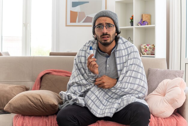 scared young ill man in optical glasses wrapped in plaid wearing winter hat holding thermometer sitting on couch at living room