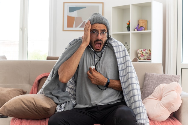 scared young ill man in optical glasses wrapped in plaid wearing winter hat measuring his pulses with stethoscope and putting hand on face sitting on couch at living room