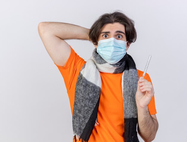 温度計を保持し、白い背景で隔離の頭の後ろに手を置くスカーフと医療マスクを身に着けている怖い若い病気の男