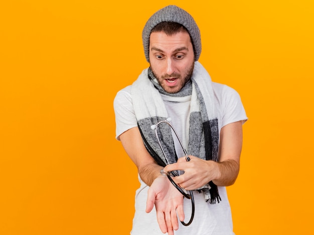 Scared young ill man wearing winter hat and scarf holding and putting stethoscope on arm isolated on yellow