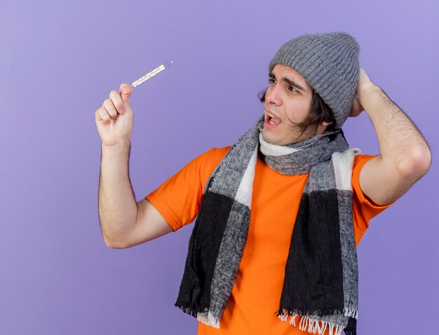 Испуганный молодой больной человек в зимней шапке с шарфом, держащий и смотрящий на термометр, положив руку на шляпу, изолированную на фиолетовом фоне
