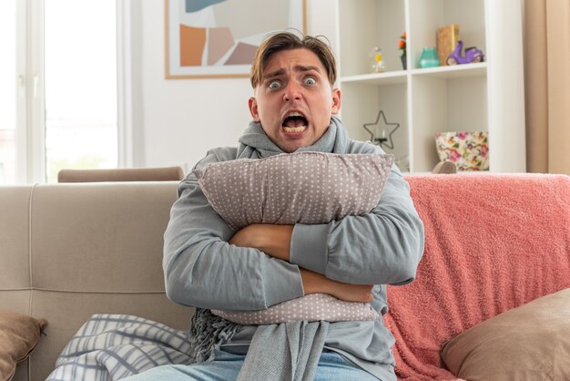 scared young ill man with scarf around neck hugging pillow sitting on couch at living room