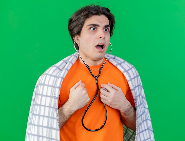 Испуганный молодой больной человек, завернутый в плед, одетый и слушающий собственное сердцебиение со стетоскопом, изолированным на зеленом