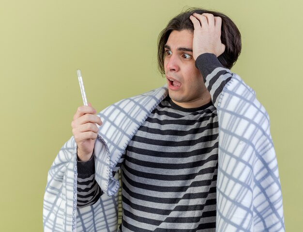 Scared young ill man wrapped in plaid holding and looking at thermometer putting hand on head isolated on olive green background