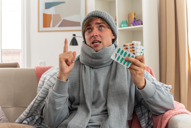 Scared young ill slavic man with scarf around neck wearing winter hat holding medicine blister packs and pointing up sitting on couch at living room