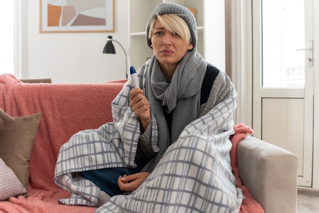 Scared young ill slavic woman with scarf around her neck wrapped in plaid wearing winter hat holding thermometer sitting on couch at living room