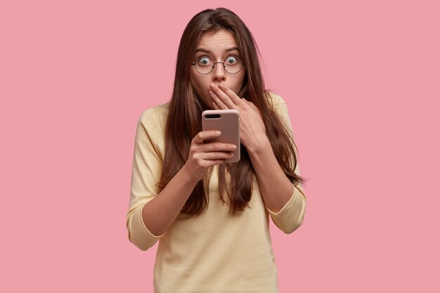scared young lady covers mouth, feels fear, carries modern cell phone, wears spectacles, reads something astonishing in internet