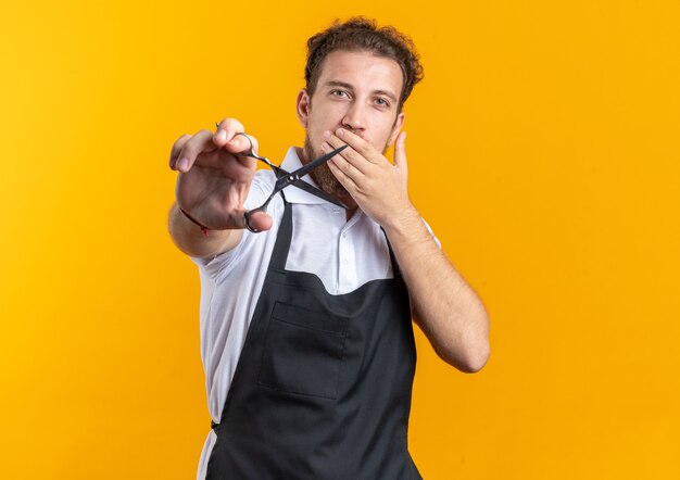 Scared young male barber wearing uniform holding out scissors at camera isolated on yellow background