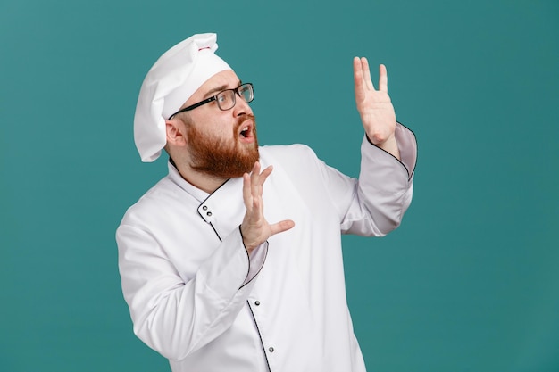 안경 유니폼을 입고 모자를 쓴 겁 먹은 젊은 남성 요리사는 파란색 배경에 고립 된 측면에 빈 손을 보여주는 측면