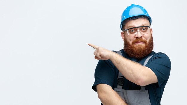 Scared young male construction worker wearing safety helmet uniform and safety glasses looking at camera pointing to side isolated on white background with copy space
