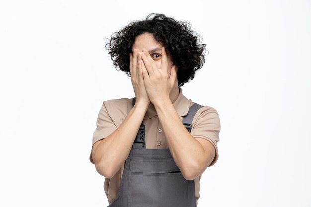 Scared young male construction worker wearing uniform closing face with hands peeking at camera through fingers isolated on white background