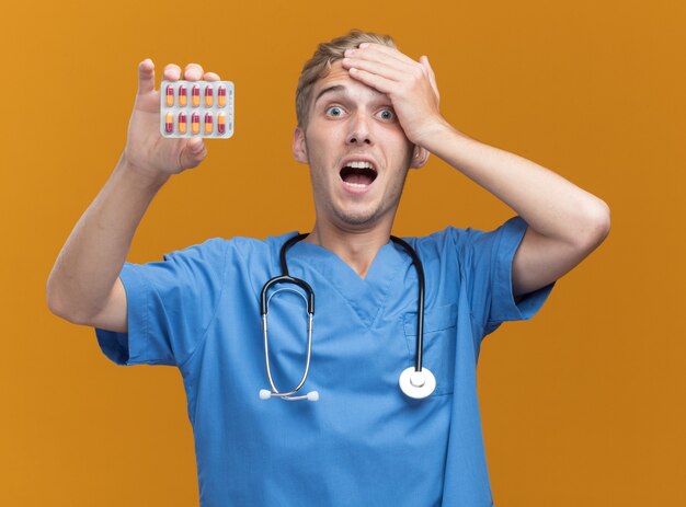 Scared young male doctor wearing doctor uniform with stethoscope holding pills putting hand on head isolated on orange wall