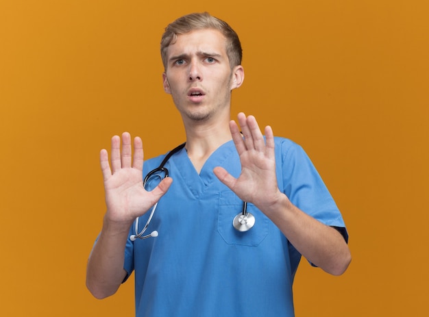 オレンジ色の壁に分離された聴診器と医師の制服を着て怖い若い男性医師