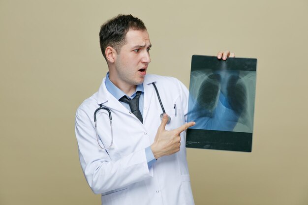 scared young male doctor wearing medical robe and stethoscope around neck showing xray shot pointing at it looking at it isolated on olive green background