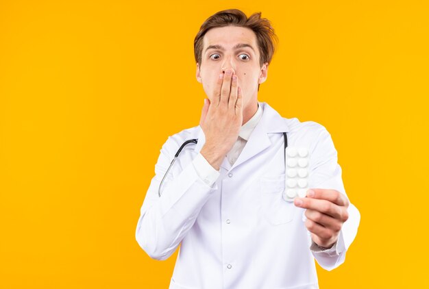 Scared young male doctor wearing medical robe with stethoscope holding and looking at pills covered mouth with hand isolated on orange wall