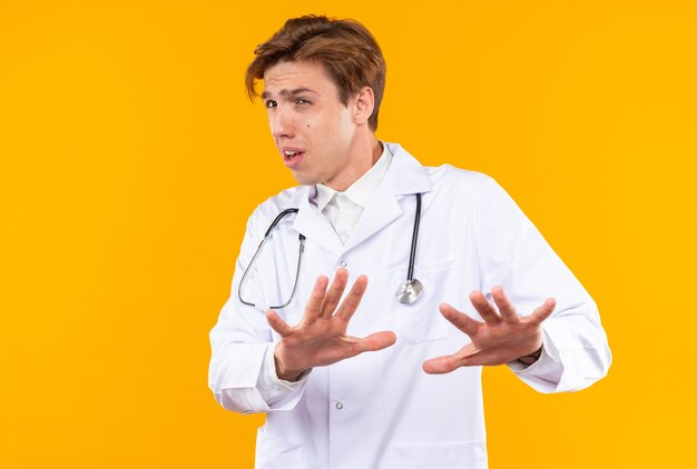 Scared young male doctor wearing medical robe with stethoscope showing stop gesture