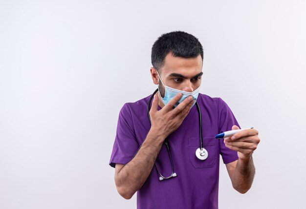 Scared young male doctor wearing purple surgeon clothing and stethoscope medical mask looking at termometer in his hand covered mouth with hand on isolated white background