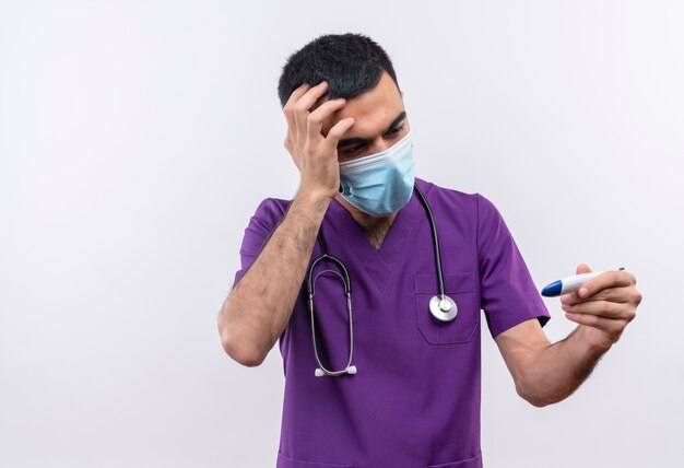 Scared young male doctor wearing purple surgeon clothing and stethoscope medical mask put his hand on forehead looking to thermometer in hand on isolated white background