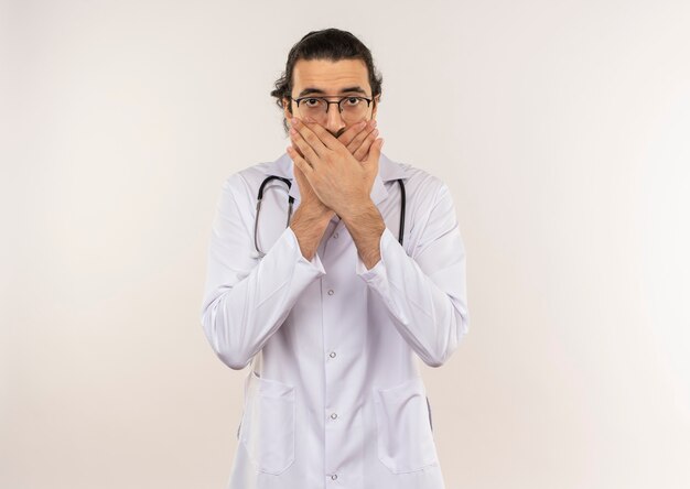 scared young male doctor with optical glasses wearing white robe with stethoscope covered mouth with hands