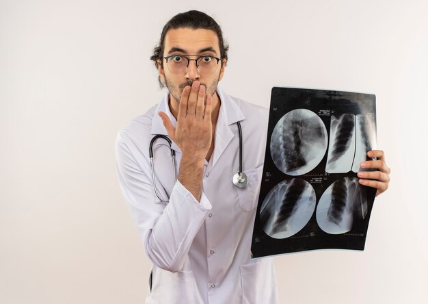 Scared young male doctor with optical glasses wearing white robe with stethoscope holding x-ray and covered mouth with hand on isolated white wall with copy space