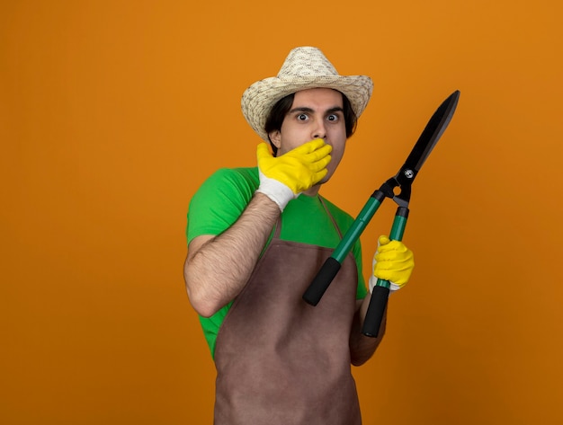 Бесплатное фото Испуганный молодой садовник в униформе в садовой шляпе с перчатками, держащими ножницы, прикрыл рот рукой