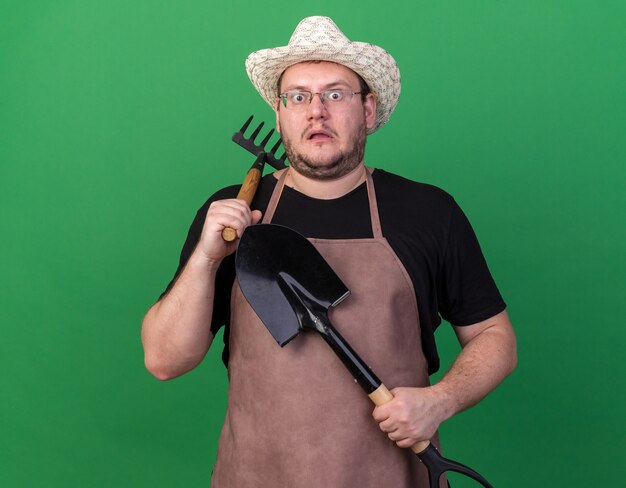 Scared young male gardener wearing gardening hat holding spade putting rake on shoulder isolated on green wall