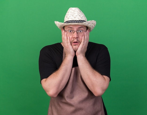 Scared young male gardener wearing gardening hat putting hands on cheeks isolated on green wall