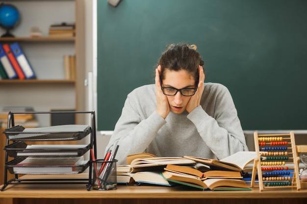 scared young male teacher grabbed head sitting at desk with school tools on in classroom