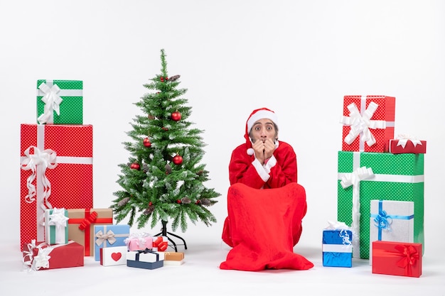 Scared young man dressed as Santa claus with gifts and decorated Christmas tree sitting on the ground on white background