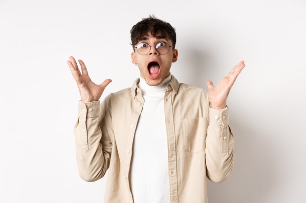 Scared young man in panic, screaming and looking axious, jumping startled and shocked, shaking hands nervously, standing on white background.