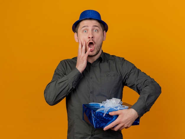 Scared young party guy wearing blue hat holding gift box putting hand on cheek isolated on orange