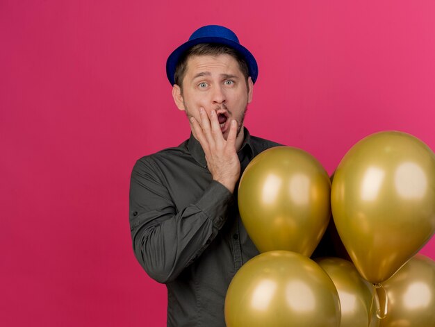 Scared young party guy wearing blue hat standing next to balloons covered mouth with hand isolated on pink