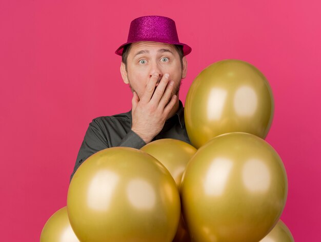 Scared young party guy wearing pink hat standing behind balloons putting and on mouth isolated on pink