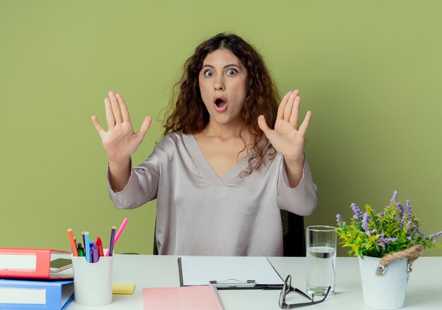 Испуганная молодая симпатичная женщина-офисный работник сидит за столом с офисными инструментами, протягивая руки к камере, изолированной на оливковом фоне