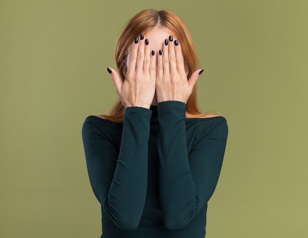 Scared young redhead ginger girl with freckles covers face with hands isolated on olive green wall with copy space