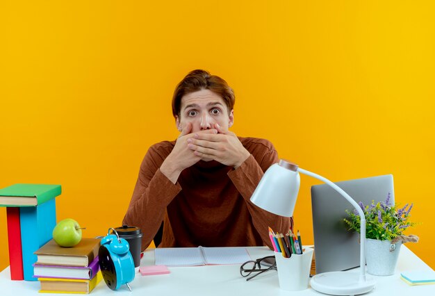 Scared young student boy sitting at desk with school tools covered mouth with hand on yellow