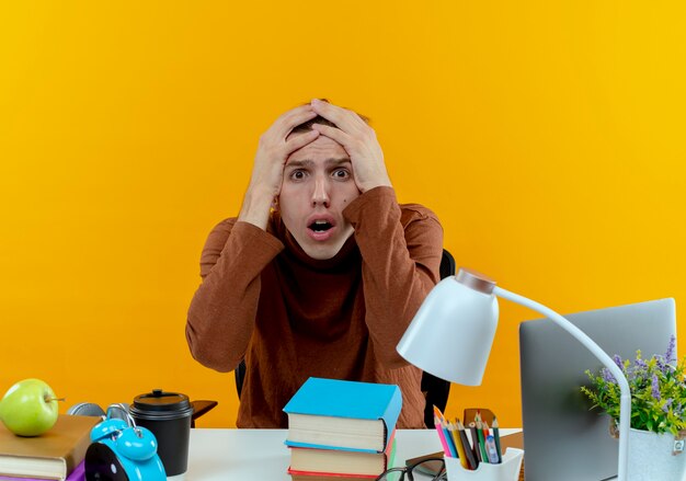 Scared young student boy sitting at desk with school tools grabbed head isolated on yellow wall