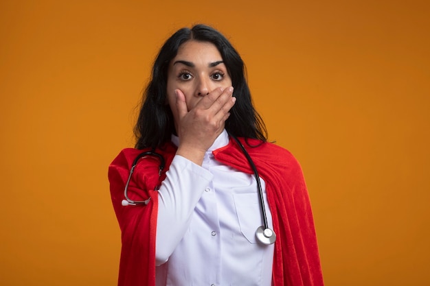 Scared young superhero girl wearing medical robe with stethoscope covered mouth with hand isolated on orange wall