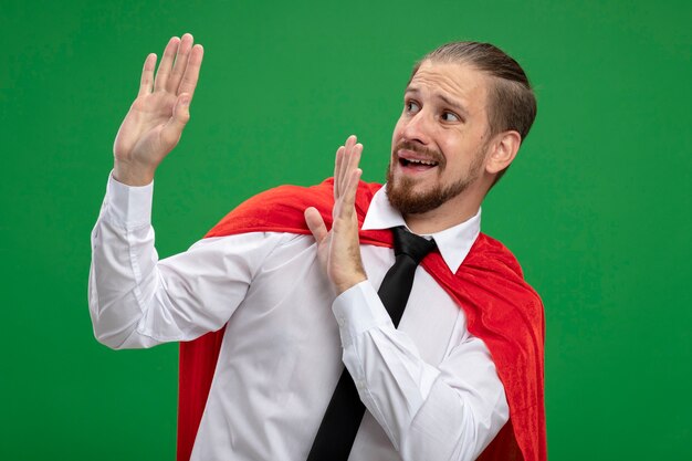 Free photo scared young superhero guy and holding out hands at side isolated on green