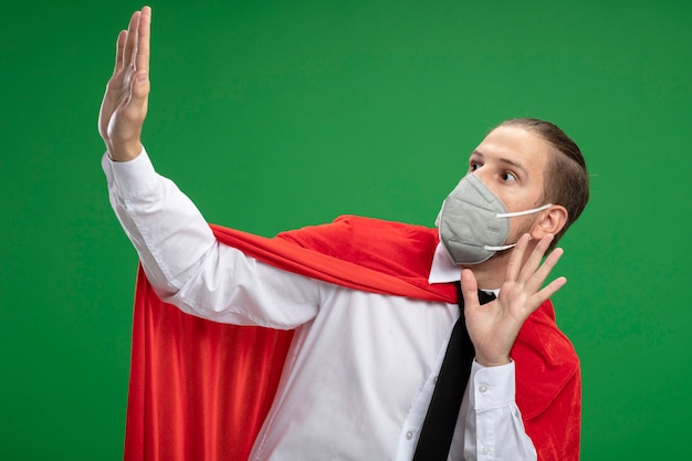 Ragazzo giovane supereroe spaventato che indossa maschera medica e cravatta guardando il lato e tendendo le mani alla macchina fotografica isolata su priorità bassa verde