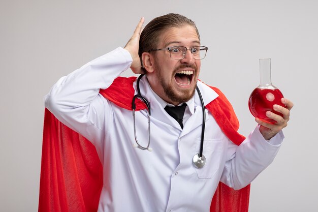 Scared young superhero guy wearing medical robe with stethoscope and glasses holding and looking at chemistry glass bottle filled with red liquid putting hand on head isolated on white