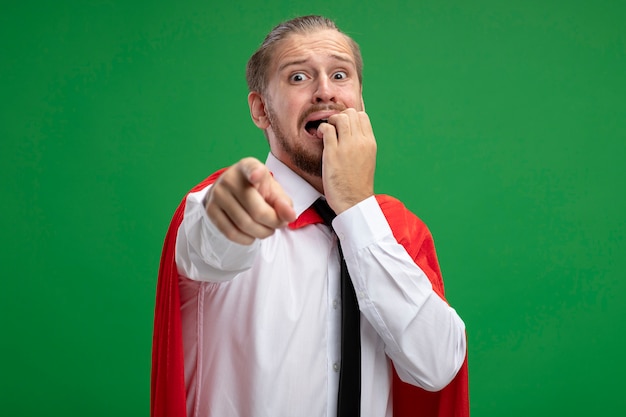 Scared young superhero guy wearing tie bites nails points at camera isolated on green background